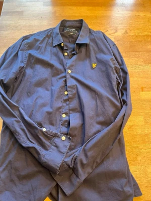 Mörkblå skjorta från Lyle & Scott - Snygg mörkblå skjorta från Lyle & Scott i slim fit-modell. Skjortan har klassisk krage, långa ärmar och vita knappar framtill. På bröstet sitter den ikoniska gula loggan med örn. Perfekt till dig som gillar stilrena plagg.