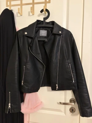 Svart skinnjacka från Lindex - Snygg svart skinnjacka från Lindex i klassisk bikerstil med silvriga dragkedjor och tryckknappar. Jackan har bred krage och detaljerade sömmar, tillverkad i konstläder. Perfekt för en cool och trendig look.