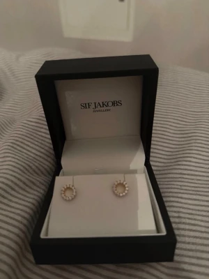 Örhängen från Sif Jakobs - Snygga örhängen från Sif Jakobs i guldton med små vita pärlor i en cirkelformad design. Perfekta för dig som gillar klassiska men ändå trendiga accessoarer. Kommer i original ask.