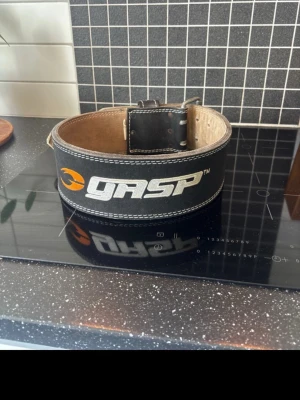 Svart träningsbälte från GASP - Säljer ett svart träningsbälte från GASP med bred design och vit kontrastsöm. Bältet har stor logga i vitt och orange framtill och är gjort i rejält skinn. Perfekt för gym och styrketräning.