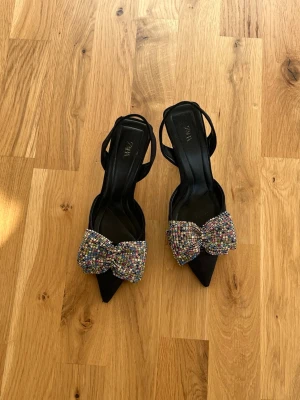 Svarta pumps med strassrosett från Zara - Säljer ett par superfina svarta klackar från Zara med spetsig tå, öppen häl & rosett på! Använda fåtal gånger har små tecken på användning där framme vid spetsen men annars som nya!  Dom är i storlek 35 men passar dig som har storlek 36! 