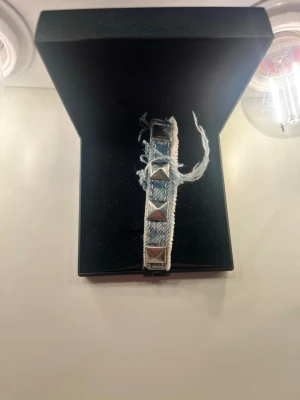 Armband i denim med nitar - Snyggt armband i ljusblå denim med slitna kanter och stora silvriga pyramidformade nitar. Armbandet har en cool och avslappnad stil, perfekt för att ge outfiten en edgy touch. Knytband i tyg för justerbar passform.