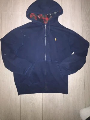 Marinblå hoodie från Polo Ralph Lauren - Snygg marinblå hoodie från Polo Ralph Lauren med dragkedja och klassisk gul logga på bröstet. Huvan har ett rutigt innerfoder i rött, grönt och vitt. Hoodien har kängurufickor och ribbade muddar. Perfekt för en chill och stilren look.