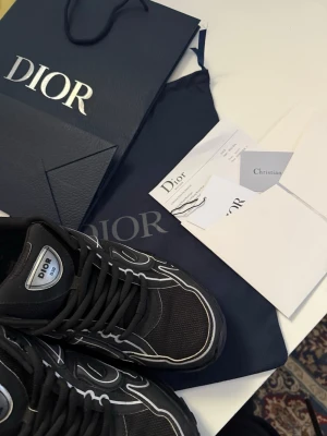 Svarta Dior sneakers med vita detaljer - Snygga svarta Dior sneakers med vita kontrastdetaljer och logga på plösen. Skorna har en sportig design med rund tå och svarta skosnören. Tillverkade i textil och läder för en exklusiv känsla. Perfekta för dig som vill ha något unikt och trendigt. Säljer mina Dior B30 sneakers i svart. Väldigt stilrena och populära sneakers från Dior.  Skick: mycket bra / nästan nya. Storlek:42  Original box, dustbag och kvitto medföljer (se bilder). Perfekta till våren och sommaren. 