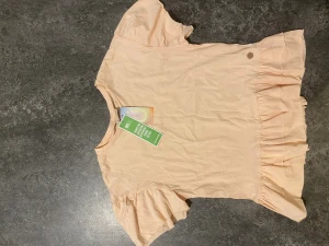 Ljusbeige volangtopp från Ü - Supersöt ljusbeige topp från Ü med korta puffärmar och volangdetalj nedtill. Tillverkad i mjuk bomull, perfekt för en trendig och avslappnad look. Enkel att matcha med jeans eller kjol.