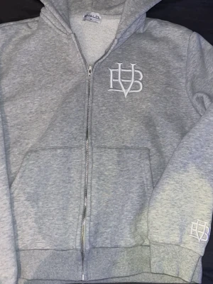 Grå zip hoodie från BVALDI - Snygg grå hoodie från BVALDI i storlek M. Dragkedja framtill, två stora fickor och broderad vit logga på bröstet och ärmen. Mjuk fleece på insidan och klassisk huva. Perfekt för chill dagar eller när du vill ha en enkel men stilren look.