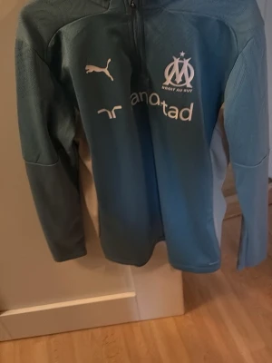 Blå Marseille träningströja Puma - Snygg blå långärmad träningströja från Puma med Olympique de Marseille-logga och sponsortryck på bröstet. Tröjan har halv dragkedja. Endast använd enstaka gånger. Pris kan sänkas vid snabb affär.