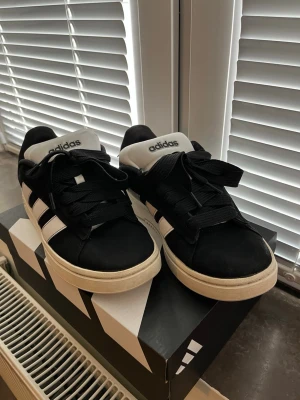 Svarta Adidas sneakers - Snygga svarta Adidas sneakers med klassiska vita ränder på sidorna och vit sula. Skorna har svart ovandel i textil eller syntet, svart snörning och vit plös med Adidas-logga. Perfekta för en sportig och stilren look.