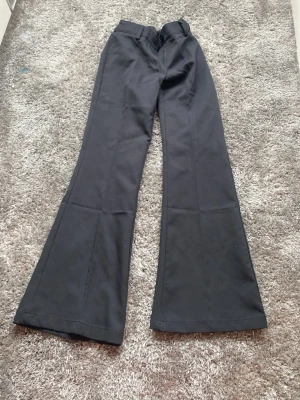 Svarta utsvängda kostymbyxor RYVLS - Snygga svarta kostymbyxor från RYVLS med utsvängda ben och klassisk passform. Byxorna har hällor för skärp och knappstängning i midjan. Perfekta för dig som gillar en trendig och stilren look.