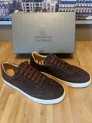 Brunello Cucinelli bruna sneakers - Mörkbruna sneakers i mocka från Brunello Cucinelli med matchande bruna skosnören och vit sula. Snygg guldfärgad logga baktill och klassisk låg modell. Perfekta för dig som gillar stilrena och exklusiva sneakers i premiumkvalitet.