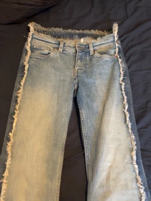 Blå jeans med fransade kanter - Unika blå jeans med fransade kanter längs sidorna och midjan. Jeansen har en klassisk femficksmodell och är tillverkade i denim med tvättad look. Perfekta för dig som vill sticka ut med en edgy stil.