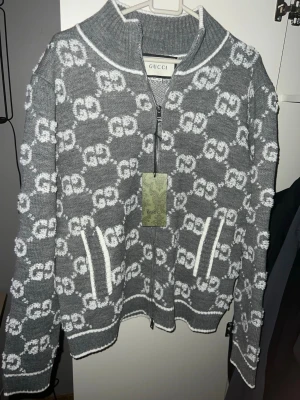 Grå stickad ziptröja från Gucci - Snygg grå stickad tröja från Gucci med vit GG-logga över hela plagget. Tröjan har halv dragkedja, två fickor framtill och ribbade muddar. Perfekt för dig som vill ha en exklusiv och trendig look. Materialet känns mjukt och lyxigt.
