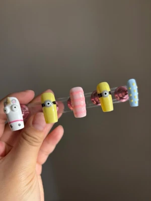 Färgglada press-on naglar med figurer - Säljer ett set press-on naglar med handmålade motiv: två gula naglar med minioner, en vit med enhörning, en rosa med ränder och en blå med små gula stjärnor. Naglarna är i plast och har en lekfull, unik design som sticker ut.
