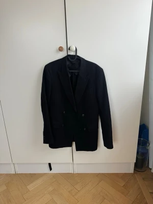 Svart dubbelknäppt kavaj från Filippa K - Stilren svart dubbelknäppt kavaj från Filippa K med dubbelknäppning och klassisk passform. Kavajen har slimmad siluett, spetsiga slag och är helfodrad med blankt innerfoder. Perfekt för en dressad look och enkel att matcha med skjorta eller t-shirt. Nypris 4000 kr 