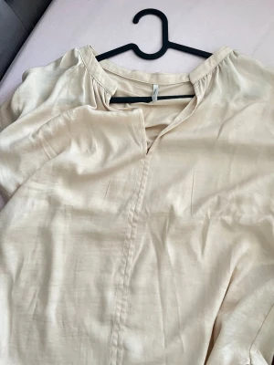 Beige blus från ONLY med v-ringning - Stilren beige blus från ONLY med v-ringad hals och korta ärmar. Tillverkad i ett mjukt, lätt tyg som ger en avslappnad känsla. Perfekt att matcha med jeans eller kjol för en enkel och snygg look.