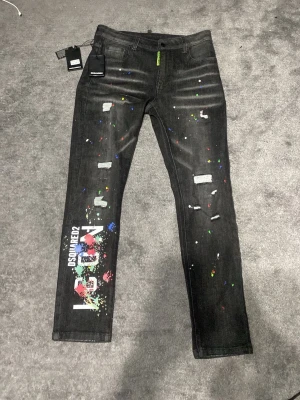 Svarta Dsquared2 jeans med färgstänk - Snygga svarta jeans från Dsquared2 med färgstänk, slitningar och tryckt text på vänster ben. Smal passform och klassisk femficksmodell. Jeansen har coola detaljer som ger en unik streetwear-look. Perfekta för dig som vill sticka ut med din stil. Pris kan diskuteras 