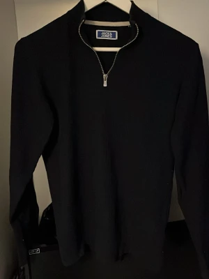 Svart halvzip-tröja Jack & Jones - Svart långärmad tröja från Jack & Jones med halv dragkedja framtill och ståkrage. Enkel och stilren design som passar perfekt till jeans eller chinos. Materialet ser ut att vara mjukt och bekvämt, perfekt för lager-på-lager.