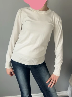 Vit stickad tröja med rund hals - Klassisk vit stickad tröja med rund halsringning och ribbade muddar vid ärmslut och nederkant. Tröjan har en enkel och stilren design som passar till det mesta. Materialet ser mjukt och bekvämt ut, perfekt för lager på lager.