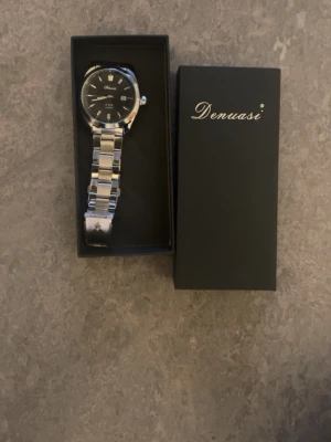 Silverfärgad klocka från Denuasi - Stilren klocka från Denuasi med svart urtavla och silverfärgat armband i metall. Klockan har tydliga markeringar och klassisk rund form. Perfekt accessoar för dig som vill ha en elegant look.