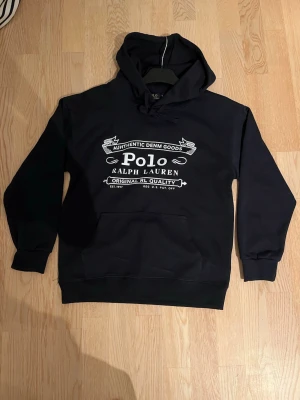 Svart hoodie från Polo Ralph Lauren - Snygg svart hoodie från Polo Ralph Lauren med stor vit logga och text på bröstet. Klassisk känguruficka framtill, ribbade muddar och justerbar huva med snören. Tillverkad i mjukt bomullsmaterial som är skönt att bära.