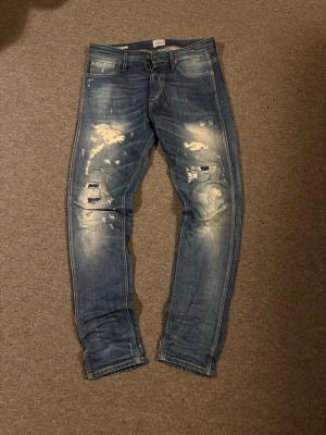 Vintage Jack & Jones jeans - Säljer ett par blå jeans med slitningar och hål på knäna. Jeansen har en slim fit passform och klassisk femficksdesign. De är tillverkade i denim och har en snygg tvättad look med ljusa partier och slitna detaljer. W29 L32, kom gärna med frågor!