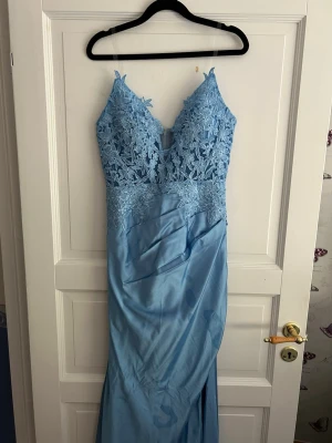 Ljusblå långklänning med spetsdetaljer - Elegant ljusblå långklänning med v-ringad spets upptill och tunna axelband. Klänningen har blommiga broderier på överdelen och en slits framtill. Perfekt för bal eller festliga tillfällen. Klänningen är endast använd 1 gång.