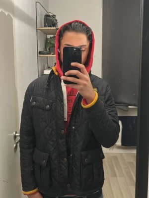 Svart quiltad Barbour jacka barn - Säljer en svart quiltad jacka från Barbour International i storlek XL barn motsvarar 170cm. Jackan har gul insida, flera fickor framtill och snygga detaljer som tryckknappar och dragkedja. Perfekt för dig som vill ha en stilren och klassisk look.