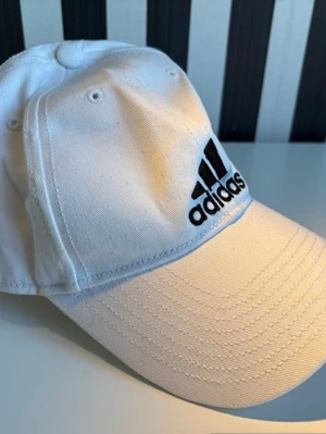 Vit keps från Adidas med logga - Snygg vit keps från Adidas med klassisk böjd skärm och svart broderad logga framtill. Kepsen är i bomullsmaterial och har ventilationshål upptill. Justerbar passform med rem bak. Perfekt för en sportig och avslappnad stil.
