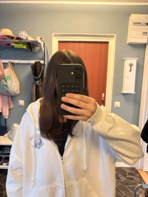 Vit hoodie med flames-tryck bak - Säljer en oversized vit hoodie med dragkedja och huva. På bröstet finns ett litet tryck och på ryggen ett stort motiv med text och flames i grått. Hoodien har snören i huvan och ribbade muddar. Perfekt för dig som gillar streetwear vibes. Kommer ej till användning.