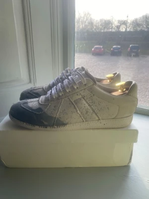 Maison margiela gats splatter - Säljer ett par vita sneakers med grå detaljer och vita skosnören. Skorna har en klassisk låg siluett och är tillverkade i canvasmaterial. Perfekta för dig som gillar en clean och stilren look med en twist.