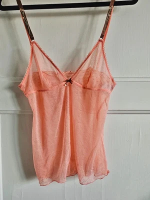 Korall färgad meshlinne med rosett - Säljer detta fantastiska meshlinne i en otrolig korall färg! 💗Toppen har tunna axelband och små prickar i tyget. Toppen har en söt svart rosett framtill och v-ringad urringning. Parfekt till sommaren! 🩷💕 är osäker på om jag vill sälja, därav priset. ställ gärna frågor! Och kika gärna på mina andra annonser! Blir rabatt vid köp av flera varor! 