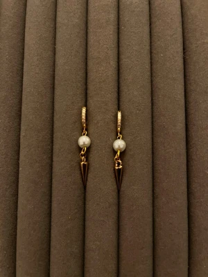 Guldörhängen med pärlor och koner - Snygga örhängen i guldton med vita pärlor och hängande konformade detaljer. Örhängena har små gnistrande stenar på bygeln och en modern, edgy design som sticker ut. Perfekta för dig som vill ha något unikt och trendigt i smyckeskrinet.