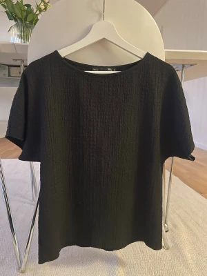 Svart strukturerad topp från KappAhl - En svart topp från KappAhl med strukturerat tyg och rund halsringning. Toppen har korta ärmar och en rak, avslappnad passform. Perfekt att styla med jeans eller kjol för en clean look.