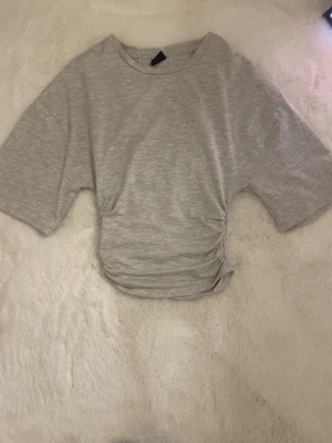 Grå croppad topp från Gina Tricot - Snygg ljusgrå croppad topp från Gina Tricot med korta ärmar och cool snörningsdetalj på sidan. Tillverkad i mjuk bomull som känns skön mot huden. Perfekt till jeans eller kjol för en chill look.