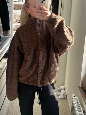 Brun oversized teddyjacka - Mysig och oversized brun teddyjacka med dragkedja och knappar framtill. Jackan har en hög krage och är tillverkad i fluffigt teddy-material som håller dig varm. Perfekt för att skapa en chill och trendig look under kalla dagar.