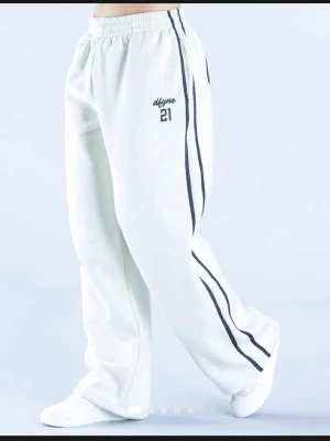Vita mjukisbyxor med svarta ränder från DFYNE - Säljer ett par vita mjukisbyxor från dfyne med svarta ränder längs benen och tryck på både framsidan och baksidan. Byxorna har avslappnad passform, resår i midjan och är tillverkade i mjukt material. Perfekta för en chill och sportig look.