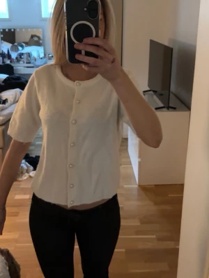 Vit kortärmad kofta med pärlknappar - Säljer en söt vit kofta med korta ärmar och & pärlknappar framtill. Koftan har en rak passform och är gjord i ett mjukt, stickat material.