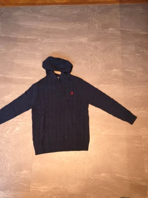 Unik hoodie från Polo Ralph Lauren - Snygg mörkblå hoodie från Polo Ralph Lauren med kabelstickat mönster och röd broderad logga på bröstet. Tröjan har huva och ribbade muddar vid ärmslut och nederkant. Perfekt för en stilren och avslappnad look. 