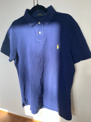Blå piké från Polo Ralph Lauren - Säljer en klassisk blå pikétröja från Polo Ralph Lauren i modellen Custom Slim Fit. Tröjan har korta ärmar, krage och två knappar framtill samt den ikoniska gula loggan broderad på bröstet. Tillverkad i bomull för en skön känsla. Storlek M.