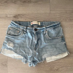 Levi's ljusblå jeansshorts med slitningar - Ljusblå jeansshorts från Levi's med rå kant och slitningar både fram och bak. Klassisk femficksmodell med knapp och dragkedja. Shortsen har låg midja och är gjorda i mjukt bomullsmaterial. 