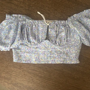 Blommig offshoulder topp med puffärm - Söt offshoulder topp med blommigt mönster i ljusa pastellfärger. Toppen har puffiga korta ärmar, snörning framtill och croppad passform. Perfekt för varma dagar. Kom gärna med prisförslag!🫶🏻