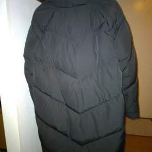 Svart längre pufferjacka / dunkappa från Mango / MNG. Strlk: S-M. Felfri. - Svart pufferjacka / dunkappa från Mango med hög krage och dragkedja framtill. Jackan har ett stilrent, quiltat mönster i V-form och stora fickor på sidorna. Perfekt för kalla dagar när du vill hålla dig varm och ändå se snygg ut. Den är i äkta dun, varm, täckande/lite längre modell. Strlk: Small, men passar eventuellt någon med Medium med. Fint skick men kan eventuellt behöva en tvätt/kemtvätt för uppfräschning. Har dock inga fläckar eller defekter. Luktar ej rök eller parfym. Ca 94 cm lång.