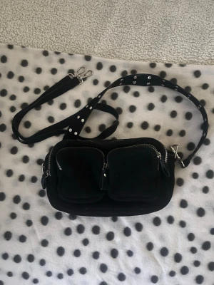 Svart axelväska med nitar och dragkedjor - Cool svart axelväska med två framfickor och silvriga dragkedjor. Väskan har en justerbar axelrem med nitar och avtagbara spännen. Perfekt för dig som gillar en edgy och praktisk look. Materialet känns mjukt och väskan har en rektangulär form. Har en liten fläck på undersidan men inget som direkt syns