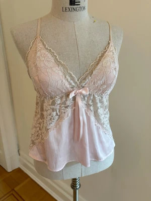 Ljusrosa spetsnattlinne med rosett - Sött ljusrosa nattlinne med tunna axelband och spetsdetaljer över bysten och ryggen. Framtill sitter en liten rosett och tyget är lätt och luftigt. Toppen har en feminin, romantisk look.