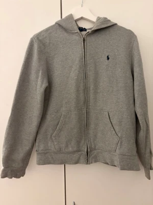Grå hoodie från Polo Ralph Lauren - Klassisk grå hoodie med dragkedja från Polo Ralph Lauren. Tröjan har huva, två fickor framtill och den ikoniska lilla mörkblå loggan broderad på bröstet. Tillverkad i mjukt sweatshirtmaterial som är skönt att ha på sig. Perfekt för en avslappnad stil.