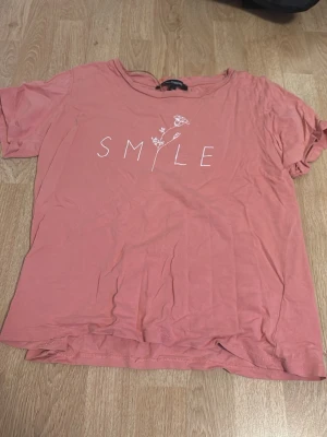 Rosa t-shirt med SMILE-tryck - T-shirt i en mjuk rosa nyans med vitt tryck där det står 'SMILE' och en liten blomdetalj. Modellen är klassisk med rund halsringning och korta ärmar. Perfekt för en avslappnad och trendig look.