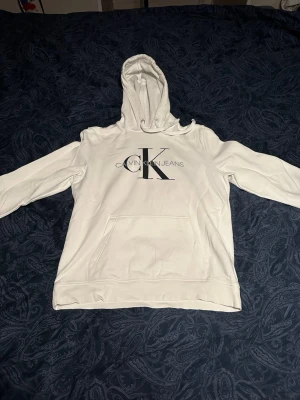Vit hoodie från Calvin Klein Jeans - Snygg vit hoodie från Calvin Klein Jeans med stor CK-logga och text på bröstet. Klassisk känguruficka framtill och justerbar huva med dragsko. Tillverkad i mjukt material som känns skönt mot huden. Perfekt för en clean och stilren look.