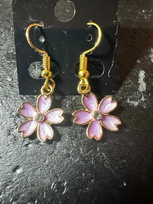 lila  blomörhängen med gulddetaljer - Söta örhängen i form av lila blommor med guldfärgade kanter och krokar. Varje blomma har en glittrande sten i mitten som ger extra bling. Perfekta för att ge din look en gullig och färgglad touch.