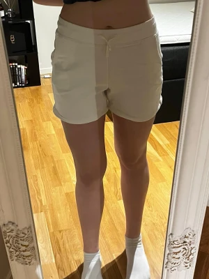 Vita shorts med snörning - Säljer ett par vita shorts med snörning i midjan och fickor på sidorna. De har en avslappnad passform och är perfekta för varma dagar. Materialet känns mjukt och bekvämt. Shortsen har en fläck på kanten, det finns en bild.