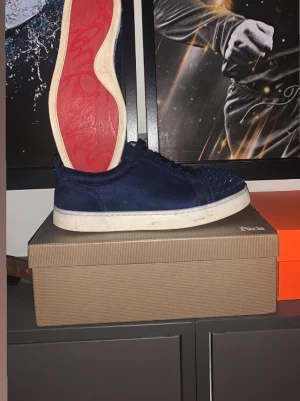 Mörkblå Christian Louboutin sneakers - Snygga mörkblå sneakers från Christian Louboutin med klassisk röd sula och nitar framtill. Skorna har lågt skaft, ovandel i mocka och vita sulor. Perfekta för dig som vill sticka ut med en lyxig och edgy stil. Saknas nån spik där fram. Pris kan diskuteras